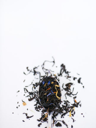 Very dark black tea blended with corn flower petals and yellow marigold petals flow over the silverspoon onto the whtie background