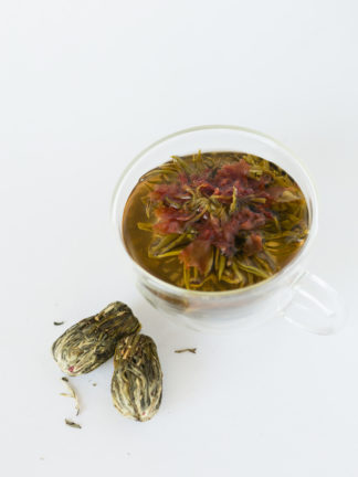 Orange carnation flower in the center of a green and white tea bouquet in the clear cup with oblong teaballs waiting to be steeped on a white background