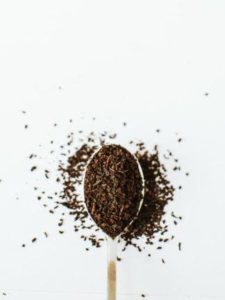 Silver tea spoon overflowing with tightly dried pieces of black tea leaves on white background