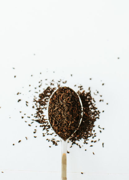 Silver tea spoon overflowing with tightly dried pieces of black tea leaves on white background
