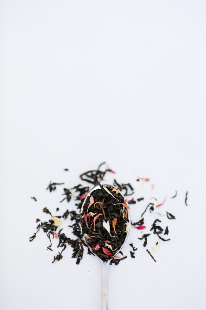 An overflowing silver spoon full of medium length black tea leaves sprinkled with a carnival of naturally colored flower petals on white background