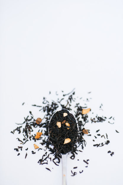 Black tea leaves and dried apricot pieces cascade over the silver spoon onto a white background