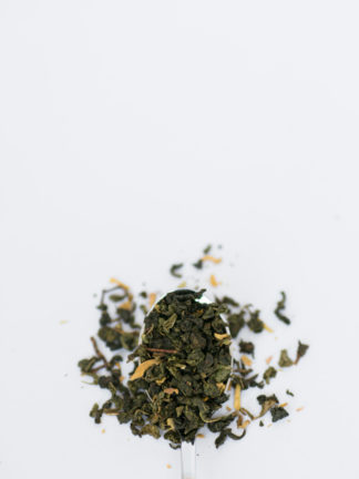 Tightly clumped rich green oolong tea leaves with truffle shavings and yellow flower petals spill over a silver spoon onto a white background