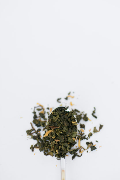 Tightly clumped rich green oolong tea leaves with truffle shavings and yellow flower petals spill over a silver spoon onto a white background