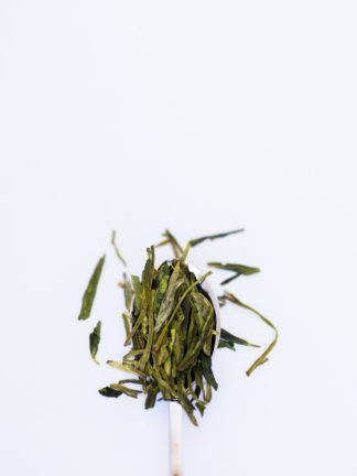 Flattened green tea leaves ranging from dark green to light green-yellow spill over the silver spoon onto the white background
