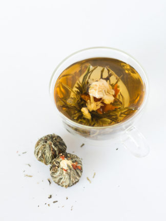 A string of jasmine flowers arch over the red lilly center surrounded by green and white teas in a clear cup with dry tea balls waiting to be steeped all on a white background