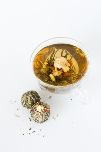 A string of jasmine flowers arch over the red lilly center surrounded by green and white teas in a clear cup with dry tea balls waiting to be steeped all on a white background
