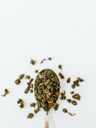 Jade green clumped oolong tea leaves resemble tea gravel spilling over the silver spoon onto the white background