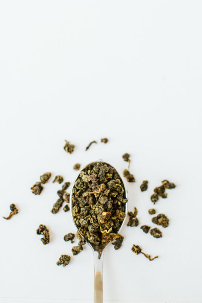 Jade green clumped oolong tea leaves resemble tea gravel spilling over the silver spoon onto the white background
