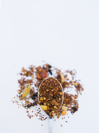 Bright orange peel pieces, cinnamon bark, and clove pieces blended in dark red rooibos overflow the silver spoon onto a white background