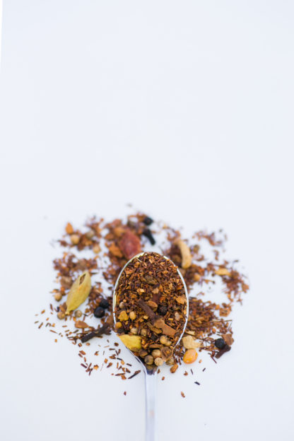 Bright orange peel pieces, cinnamon bark, and clove pieces blended in dark red rooibos overflow the silver spoon onto a white background