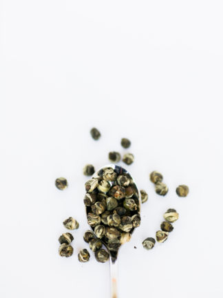 Varigated light and dark green tea leaves tightly wrapped tea pearls spill over the silver spoon onto the white background