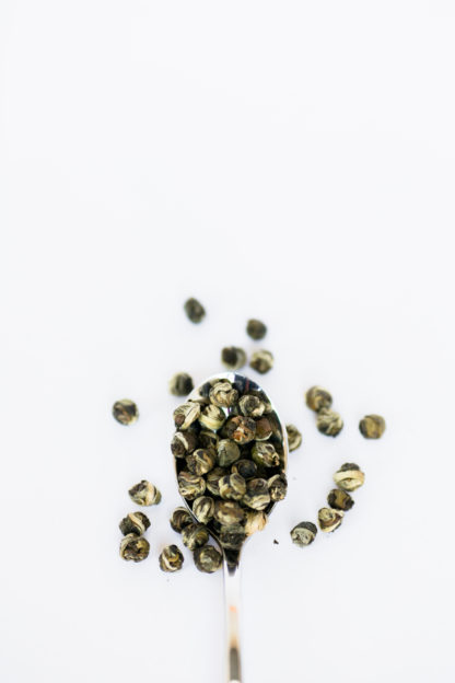 Varigated light and dark green tea leaves tightly wrapped tea pearls spill over the silver spoon onto the white background