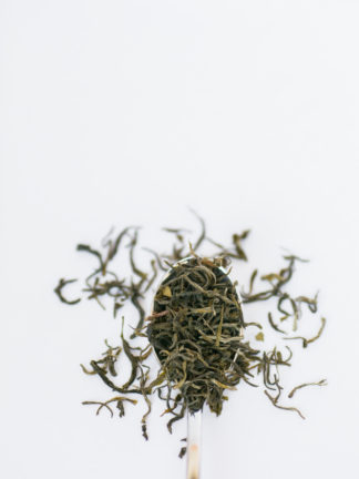 Wiry green tea leaves overflowing the silver spoon onto the white background