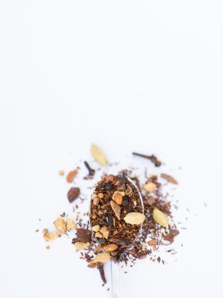 Dark red rooibos needles blended with cloves, cardamom pods, and cinnamon bark chips spill over the silver spoon onto a white background