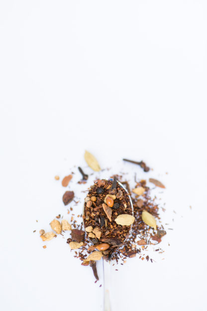 Dark red rooibos needles blended with cloves, cardamom pods, and cinnamon bark chips spill over the silver spoon onto a white background