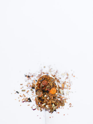 Dried orange & red rose hips, with clove and cinnamon bark chips sprinkled among dark red rooibos needles spill over the spoon onto a white background