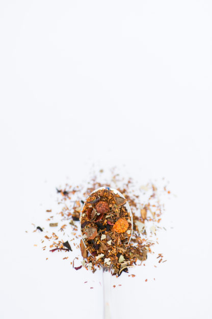Dried orange & red rose hips, with clove and cinnamon bark chips sprinkled among dark red rooibos needles spill over the spoon onto a white background