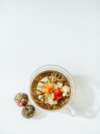 Yellow carnation and red amaranth flower and a string of white jasmine flowers unfold from the center of green and white tea wrapped tea ball in a clear glass cup