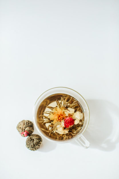 Yellow carnation and red amaranth flower and a string of white jasmine flowers unfold from the center of green and white tea wrapped tea ball in a clear glass cup