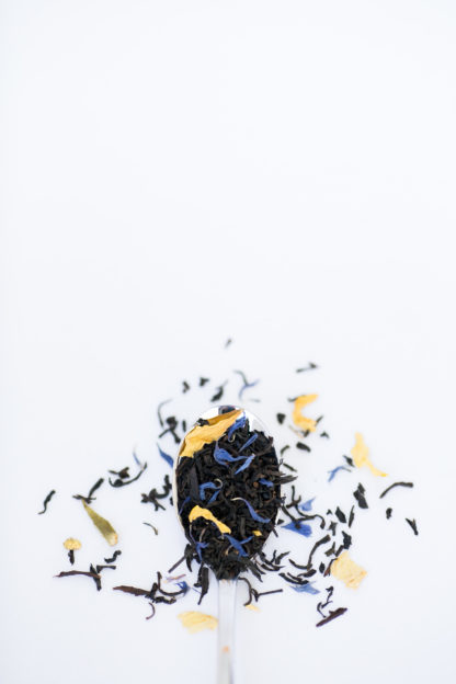 Bright yellow safflowers and dark blue corn flowers blended with dark black tea overflowing a silver spoon onto a white background