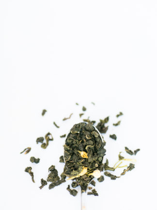 Jade green classic clumped oolong tea leaves with marigold flower petals spill over the silver spoon onto the white background