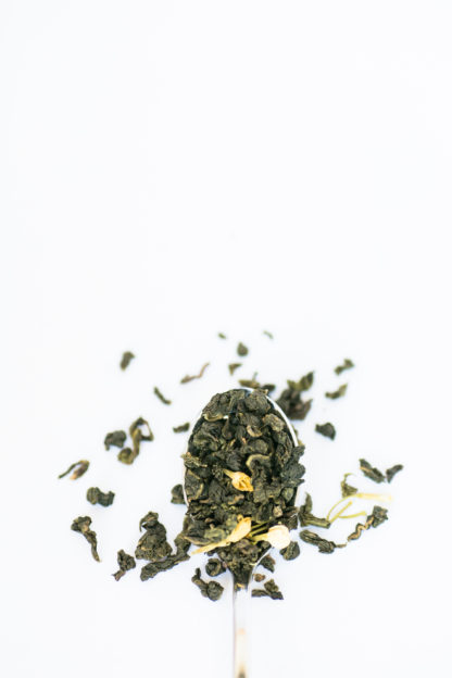 Jade green classic clumped oolong tea leaves with marigold flower petals spill over the silver spoon onto the white background