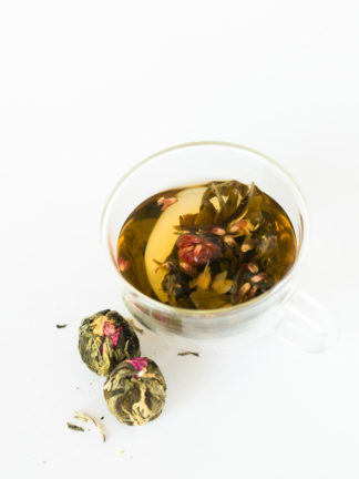 A bouquet of pink globe aramath flowers are cradled by green and white tea leaves in a clear glass cup with the dry blooming teas waiting to be steeped all on a white background