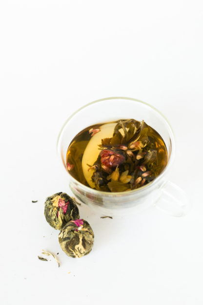 A bouquet of pink globe aramath flowers are cradled by green and white tea leaves in a clear glass cup with the dry blooming teas waiting to be steeped all on a white background