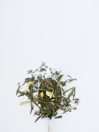 Flat dried green tea leaves are blended with jasmine flowers and orange pieces of dried mango displayed on a silver spoon overflowing onto a pure white background
