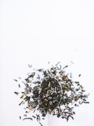 Purple lavender florets mix with dark black tea leaves spilling over the silver spoon onto the white background