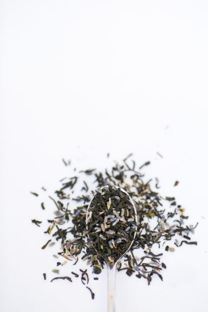 Purple lavender florets mix with dark black tea leaves spilling over the silver spoon onto the white background