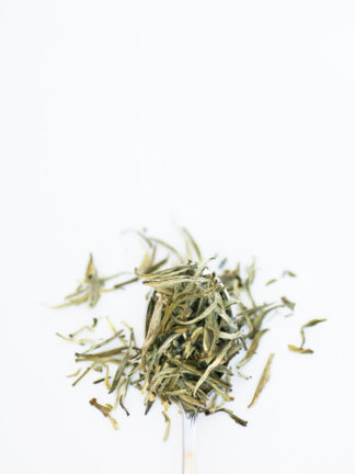 Dark blue lavender florets dot a spoonful of light green and white tea needles on a white background