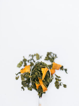 Dried yellow peach pieces are sprinkled among the dark green clumped tea leaves overflowing the silver spoon onto the white background