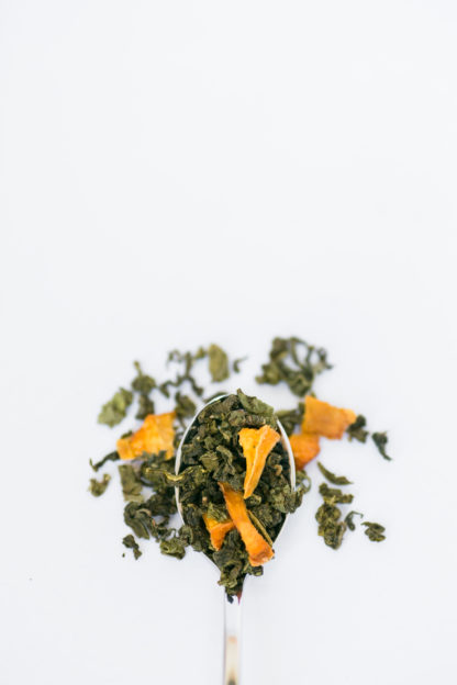 Dried yellow peach pieces are sprinkled among the dark green clumped tea leaves overflowing the silver spoon onto the white background