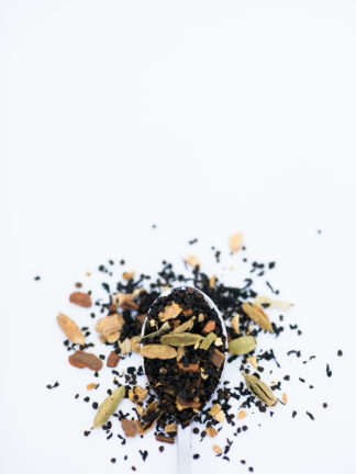 Filely chopped black tea leaves mix with green coriander pods, cloves, and rusty cinnamon bark spill onto a white background