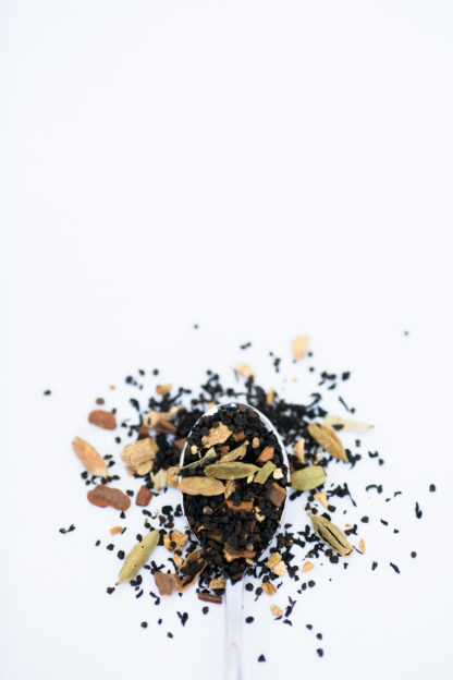 Filely chopped black tea leaves mix with green coriander pods, cloves, and rusty cinnamon bark spill onto a white background