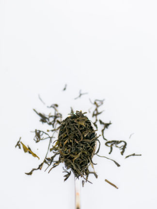 Thin dark tea leaves like twisted threads overflow the spoon onto the white background