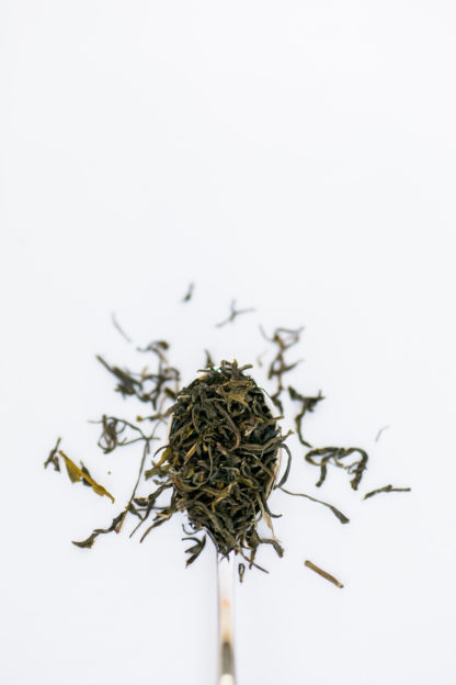 Thin dark tea leaves like twisted threads overflow the spoon onto the white background