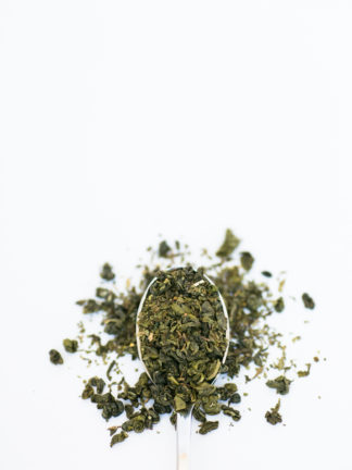 Bright green chopped mint leaves are blended with darker green classically clumped gunpowder green tea overflowing the silver spoon onto the pure white background