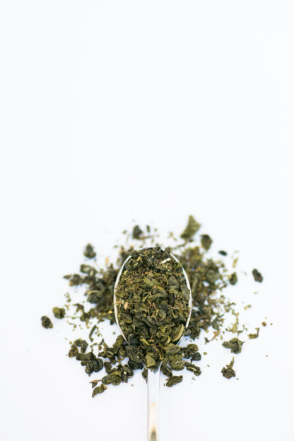 Bright green chopped mint leaves are blended with darker green classically clumped gunpowder green tea overflowing the silver spoon onto the pure white background