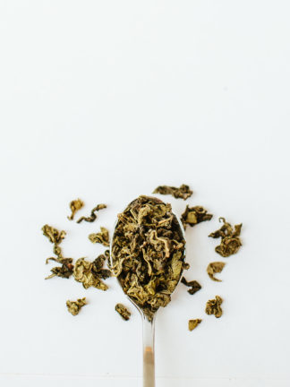 Variegated dark green and light green clumped oolong tea leaves overflow the silver spoon onto a white background