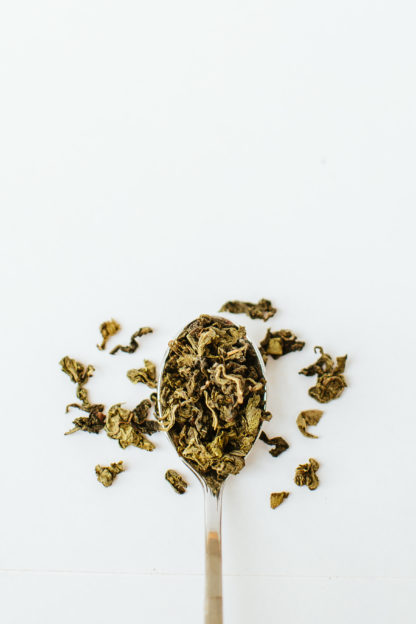 Variegated dark green and light green clumped oolong tea leaves overflow the silver spoon onto a white background