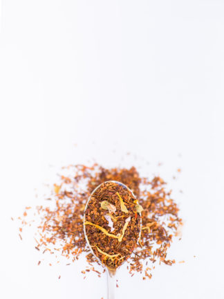 Bright yellow safflower and marigold, blue mallow flower and cornflower petals blended into dark red rooibos needles overflow a silver spoon onto a white background