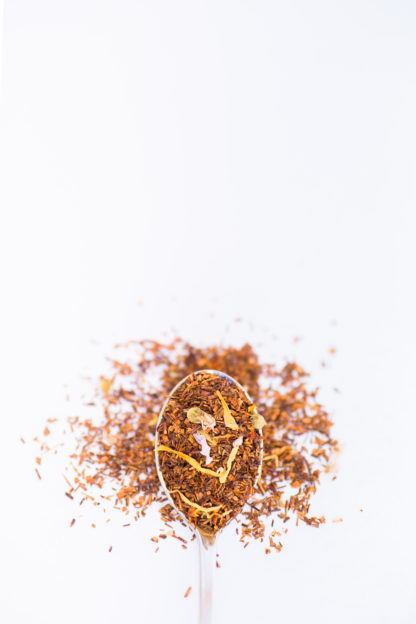 Bright yellow safflower and marigold, blue mallow flower and cornflower petals blended into dark red rooibos needles overflow a silver spoon onto a white background