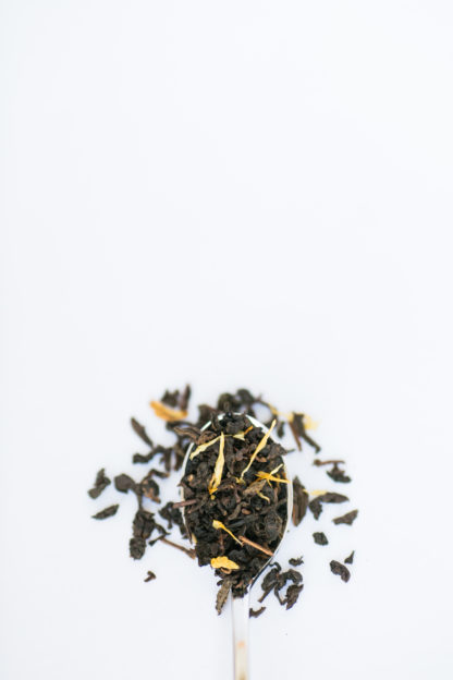 Yellow marigold flower petals are sprinkled among the clumped classic oolong tea leaves overflowing the silver spoon onto the white background
