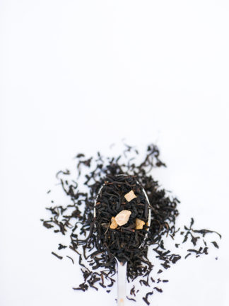 Dark brown black tea leaves dotted with dried peach pieces spill over the silver spoon onto the white background