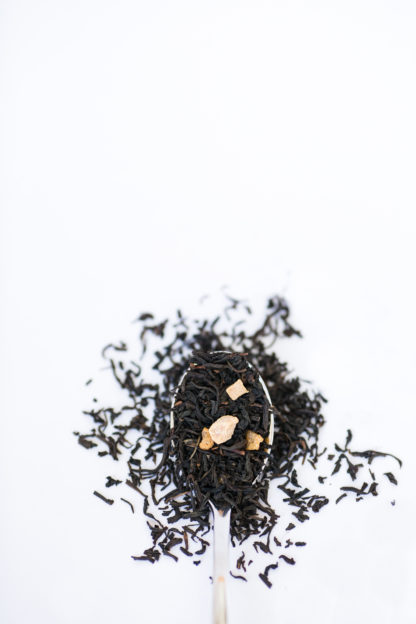 Dark brown black tea leaves dotted with dried peach pieces spill over the silver spoon onto the white background