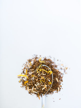 Bright yellow shredded dried peach pieces blend with dark red chopped rooibos needles overflow onto white background