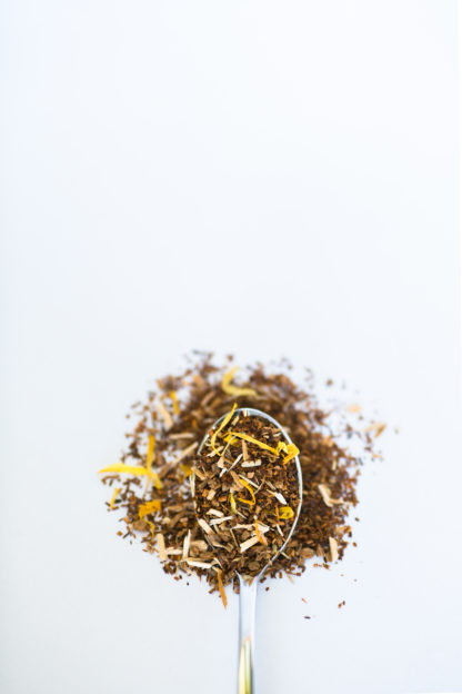 Bright yellow shredded dried peach pieces blend with dark red chopped rooibos needles overflow onto white background